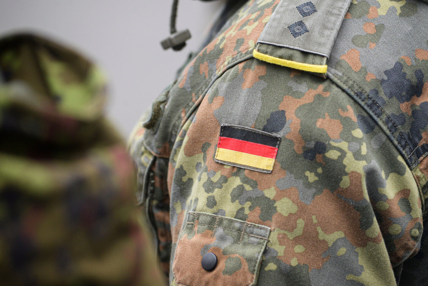 A German soldier is wearing military gear. On their left shoulder is a patch of the German flag.