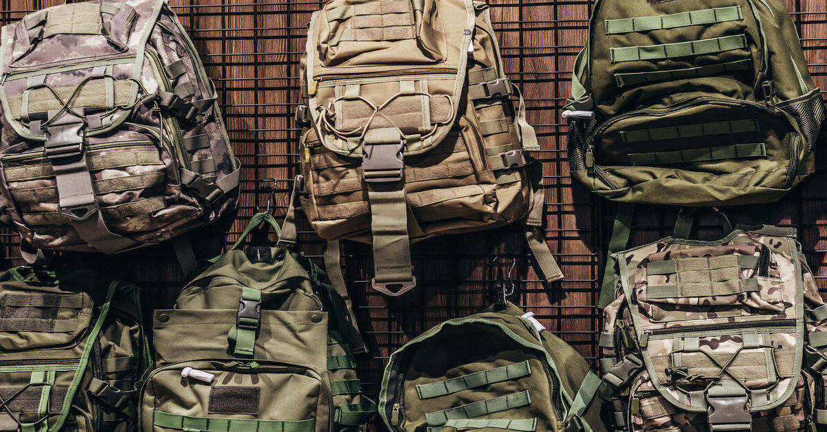 A metal rack against a wood wall with a selection of various military rucksacks hung on hooks. A metal rack against a wood wall with a selection of various military rucksacks hung on hooks.