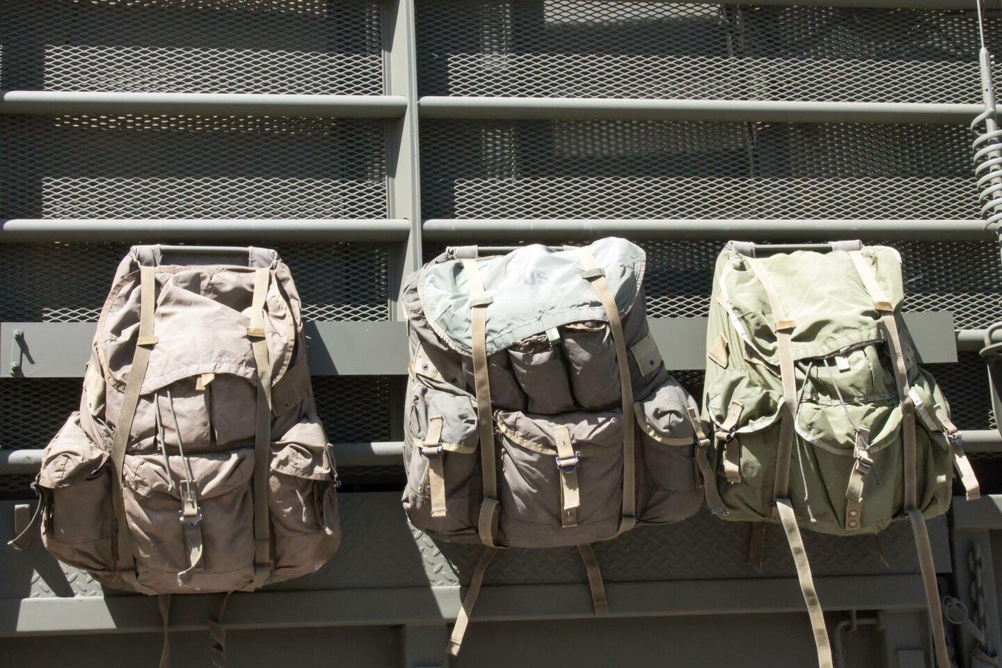 Three military rucksacks, in varying sizes and colors, are hanging on the back of a military truck.