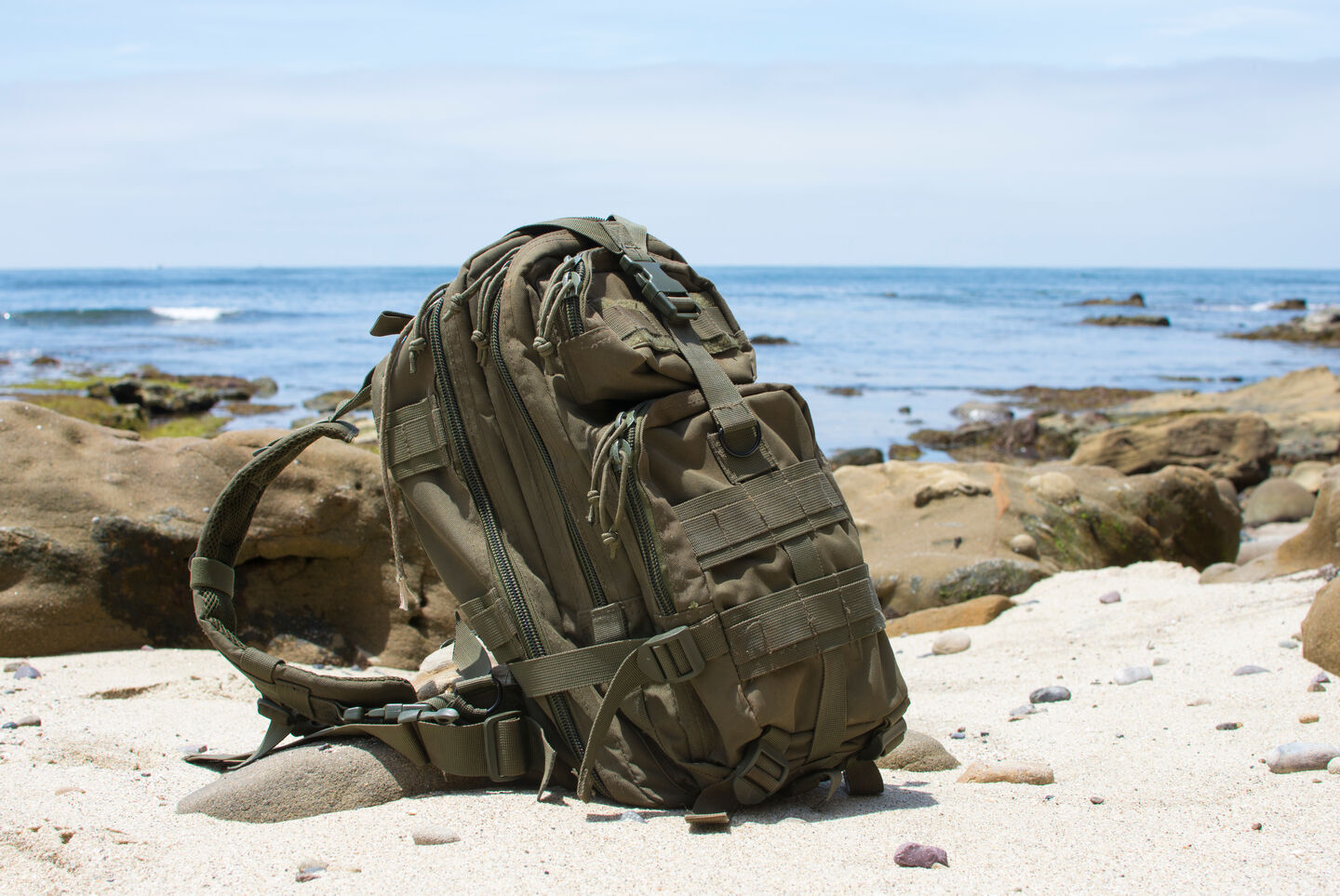 A military ruck backpack sitting on the sandy beach with ocean rolling in in the background, on a sunny day.