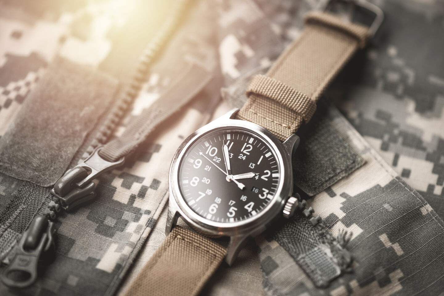 A close-up of a silver, chunky wristwatch on top of a camouflage jacket. A light source glints, reflecting off the metal surface of the watch. A close-up of a silver, chunky wristwatch on top of a camouflage jacket. A light source glints, reflecting off the metal surface of the watch.